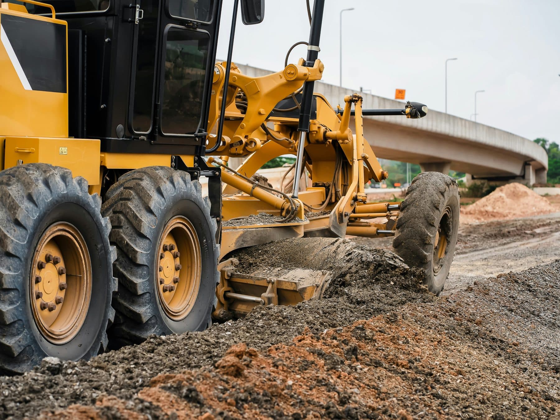 A motor grader trimming a road formation close to a completed concrete bridge structure.