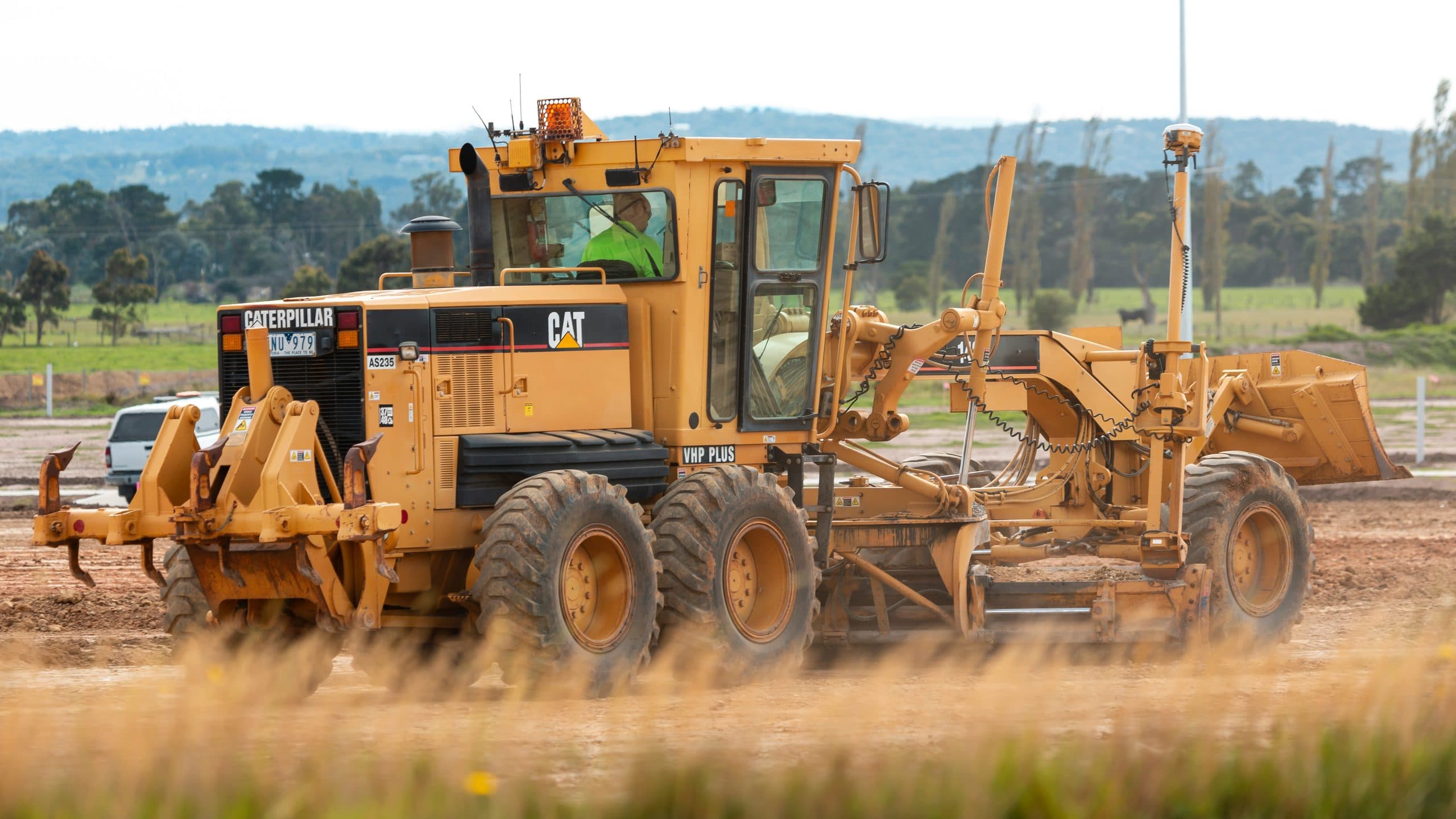 A motor grader shaping a road formation on an open site, with a compaction roller working behind it.