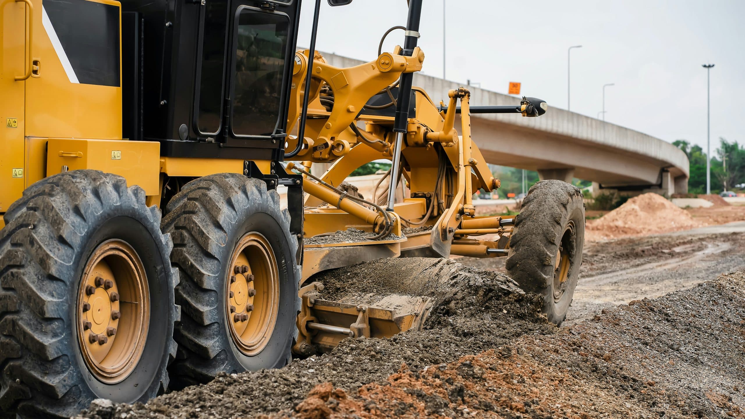 A motor grader trimming a road formation beside a completed concrete bridge structure.