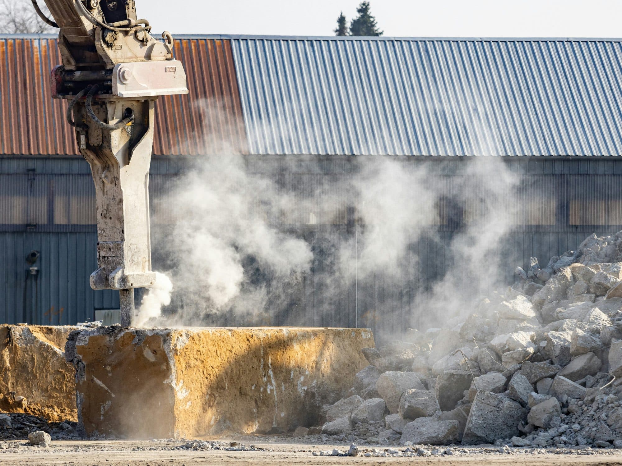 A hydraulic rock breaker mounted on an excavator arm breaking oversize rock, throwing up dust.